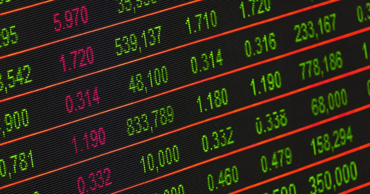 Close-up of a digital stock ticker display glowing with green, pink and orange numbers against a black background, conveying a tense financial-market mood